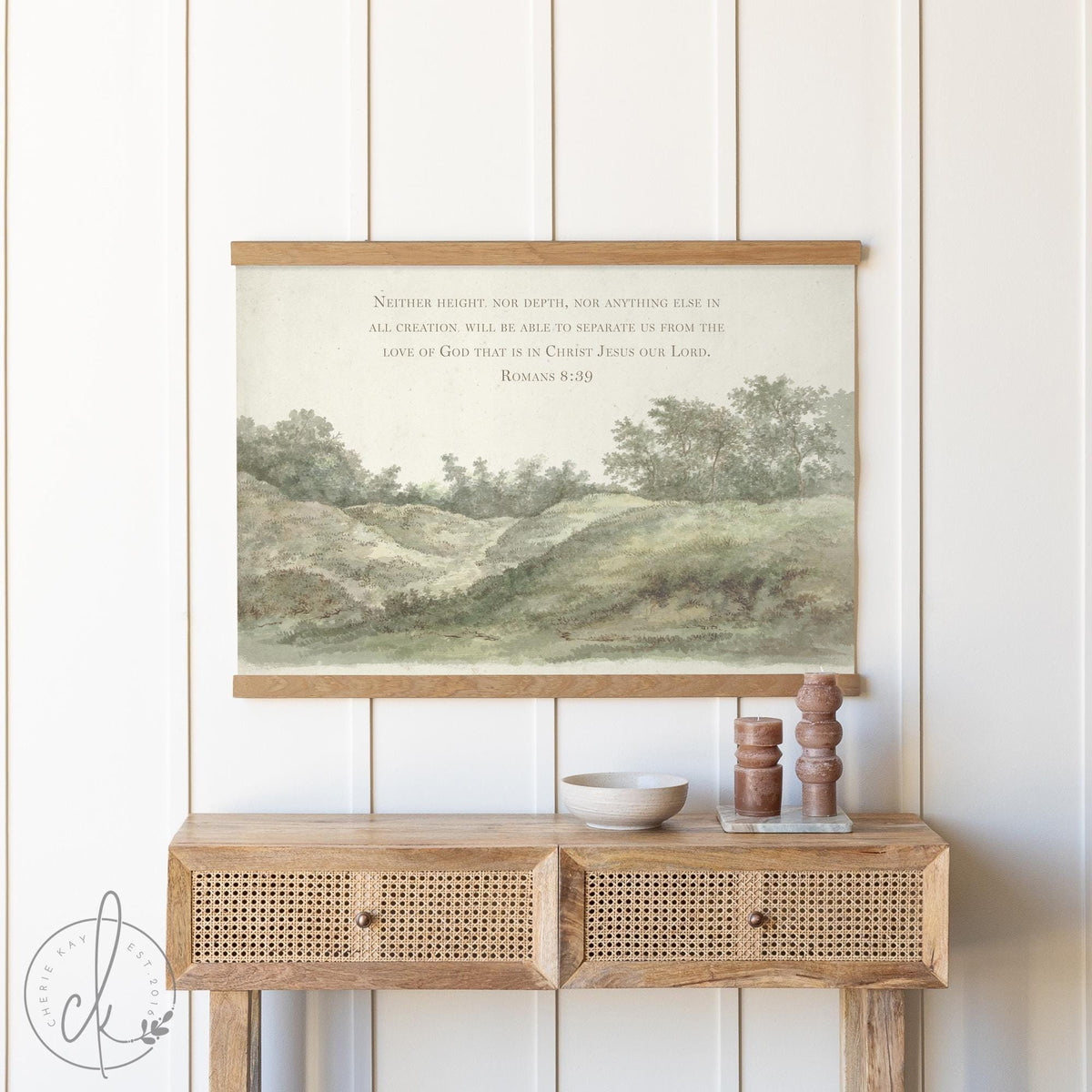 Rustic entryway decor featuring a vintage landscape wall tapestry with a countryside scene and Romans 8:39 scripture, framed by wooden accents, styled above a woven rattan console table with neutral decor items