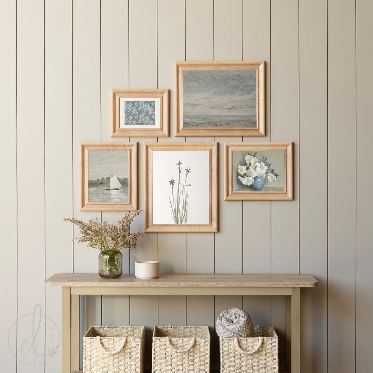 Coastal vintage gallery wall art set, including an ocean sky, a sailboat on the water, delicate wildflowers, white roses in a blue vase, and a blue floral pattern, displayed above a console table with woven baskets for a beach house or coastal decor.