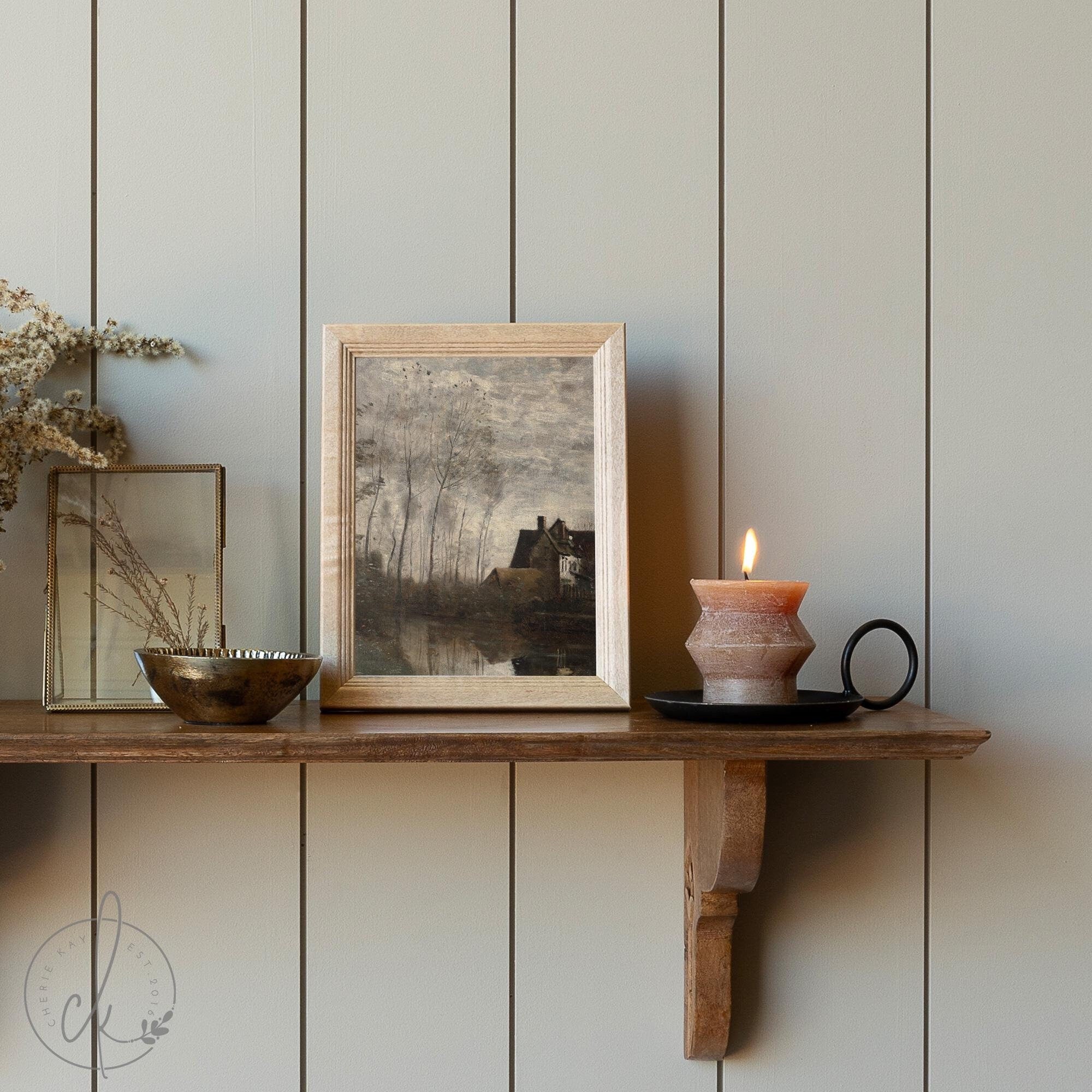 Framed vintage landscape artwork on a rustic wooden shelf, styled with a lit candle, brass bowl, and dried florals against a neutral paneled wall. Cozy and minimalist home decor aesthetic.