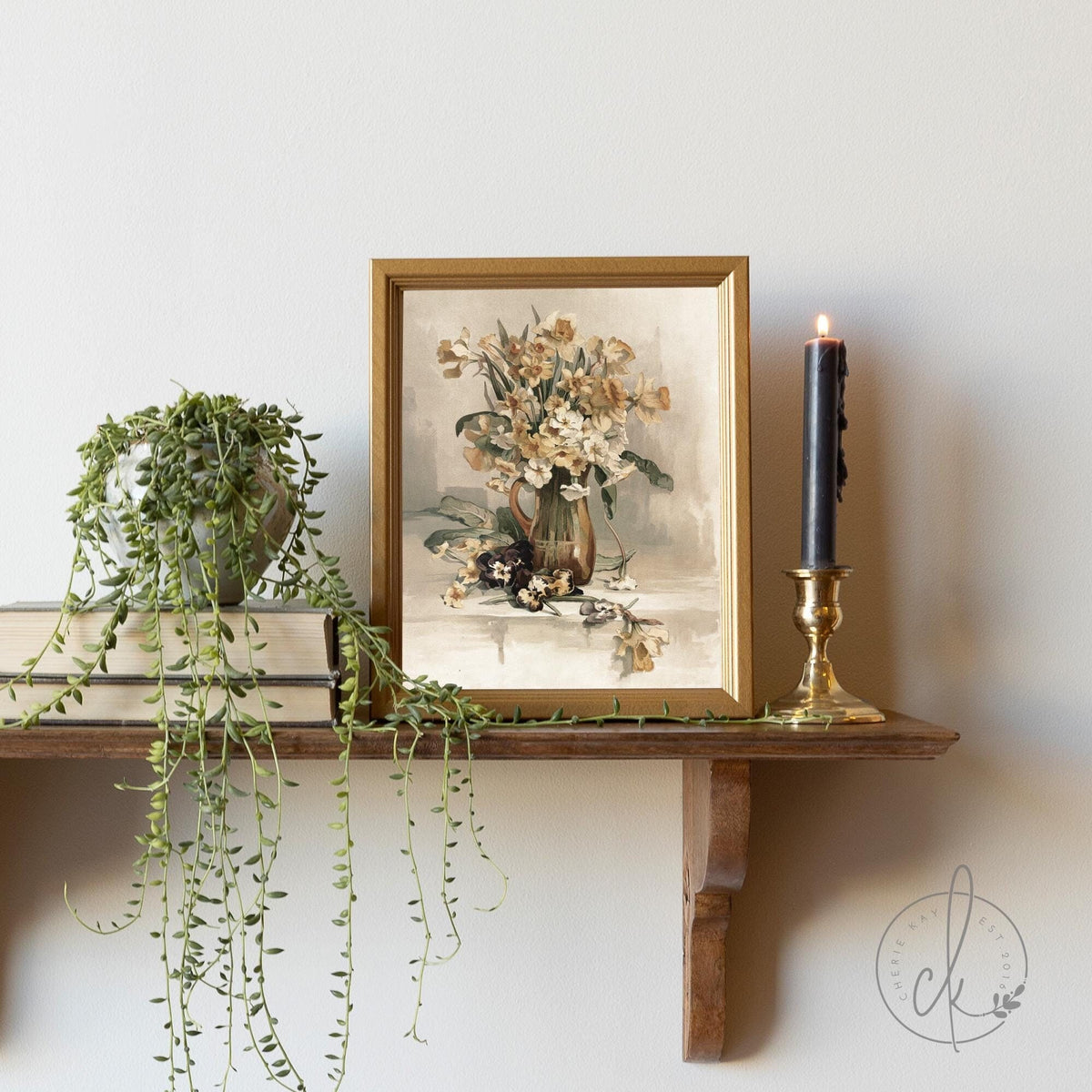 Framed vintage-style floral artwork on a wooden shelf, accompanied by a trailing green plant, stacked books, and a black candle in a brass holder.