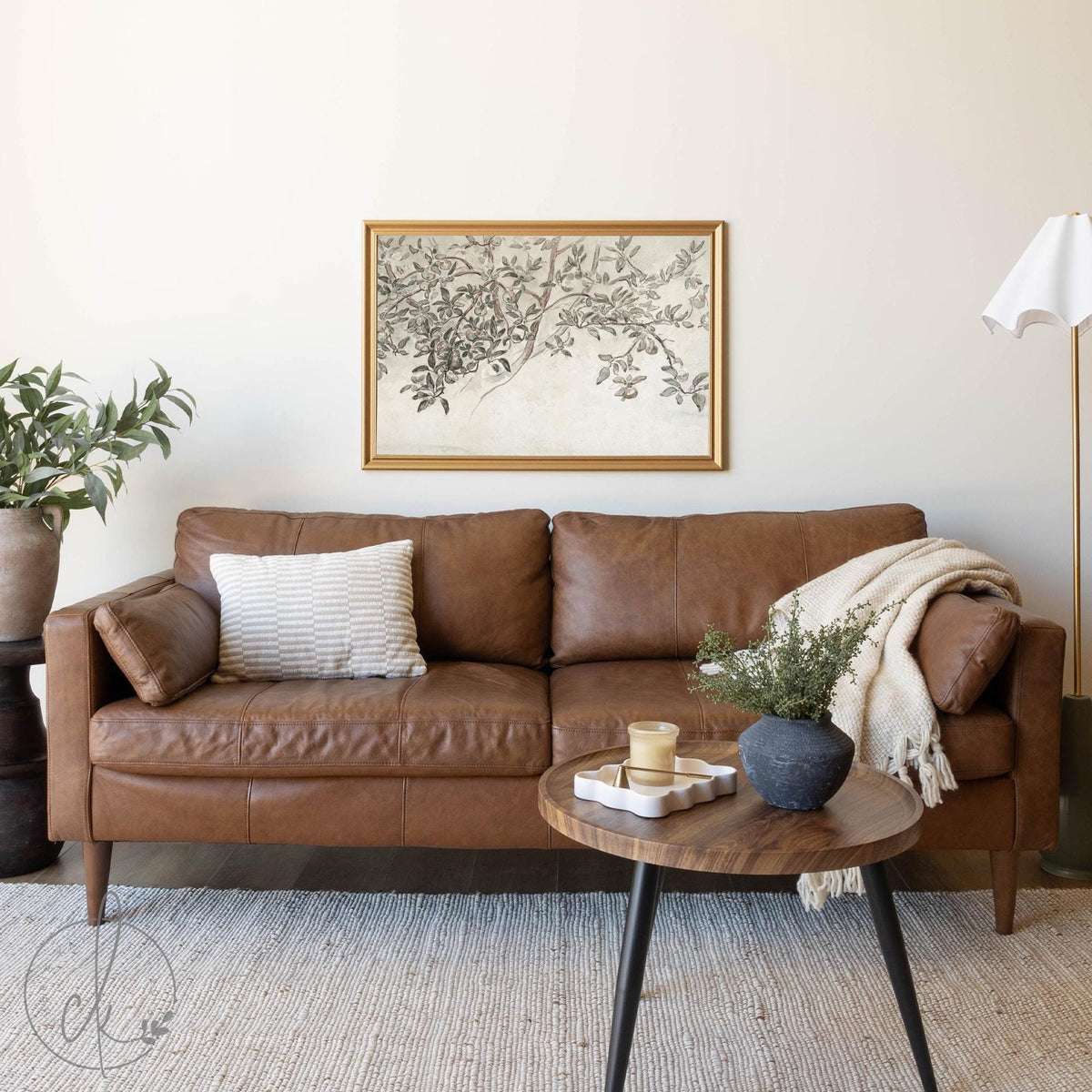 Cozy living room featuring a brown leather sofa with a neutral pillow and throw blanket, complemented by a framed botanical landscape wall art in a gold frame, a coffee table with decorative accents, and a potted plant adding a touch of greenery.