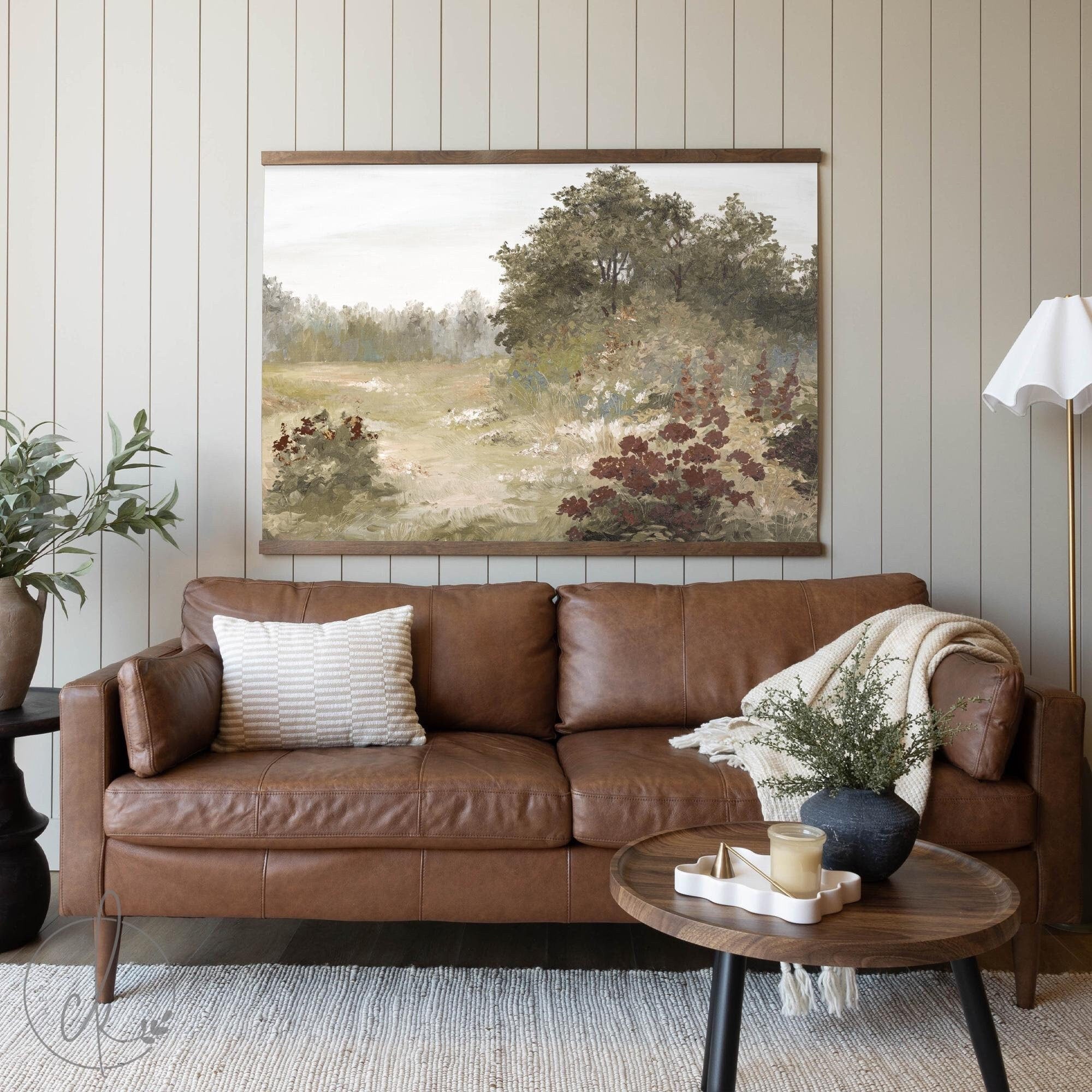 A cozy living room with a brown leather sofa, a textured pillow, and a blanket. A round wooden coffee table with decor sits in front, and a wall art piece of a vibrant meadow scene hangs above.