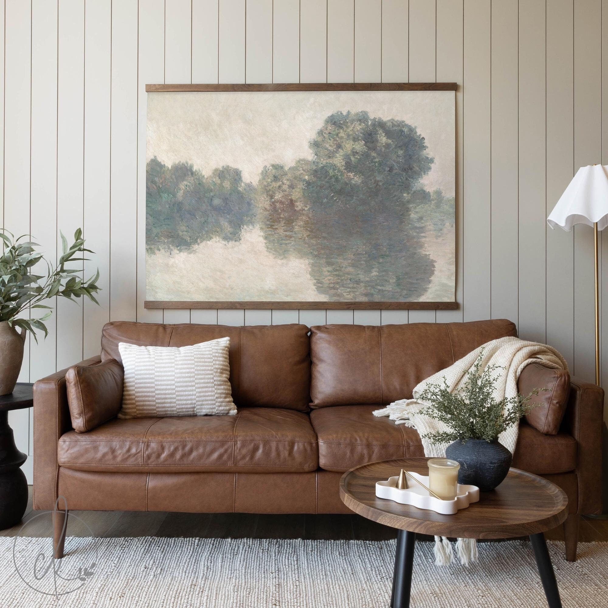 A cozy living room with a brown leather sofa, a textured pillow, and a blanket. A round wooden coffee table holds decor, and a wall-mounted art piece of a peaceful river scene hangs above.