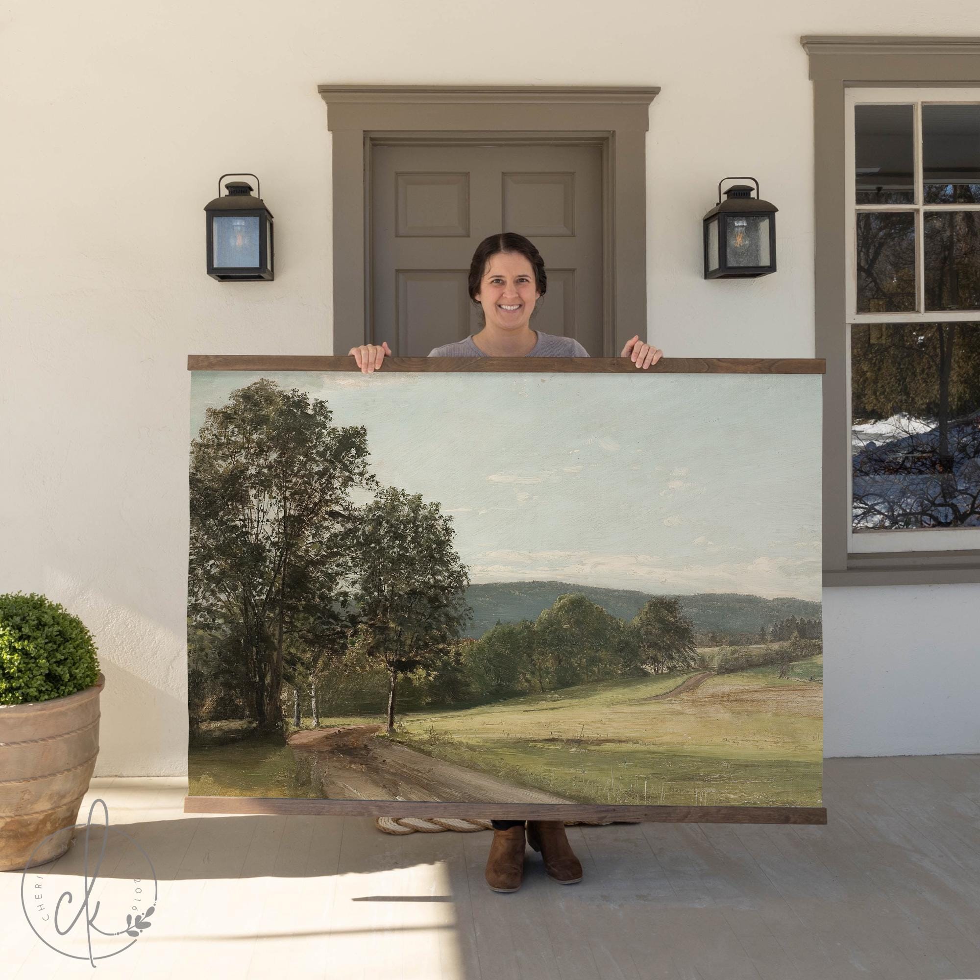 A person standing outside a house holding a large wall art piece depicting a sunny countryside scene with a dirt path, trees, and rolling hills. The background includes a door, wall-mounted lights, and a potted plant.