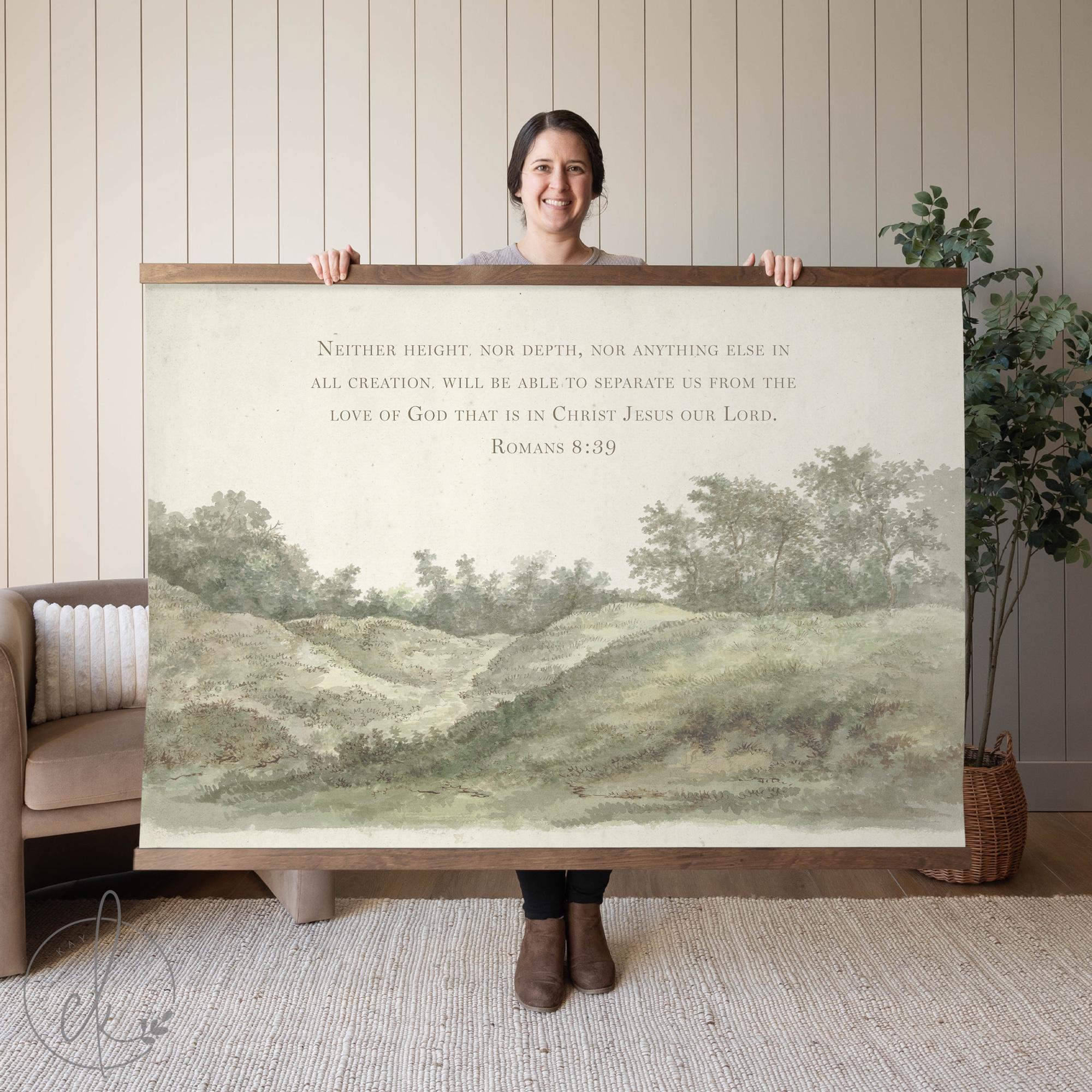 A person holding wall art of a countryside landscape with a verse from Romans 8:39. The background features a sofa and a potted plant.