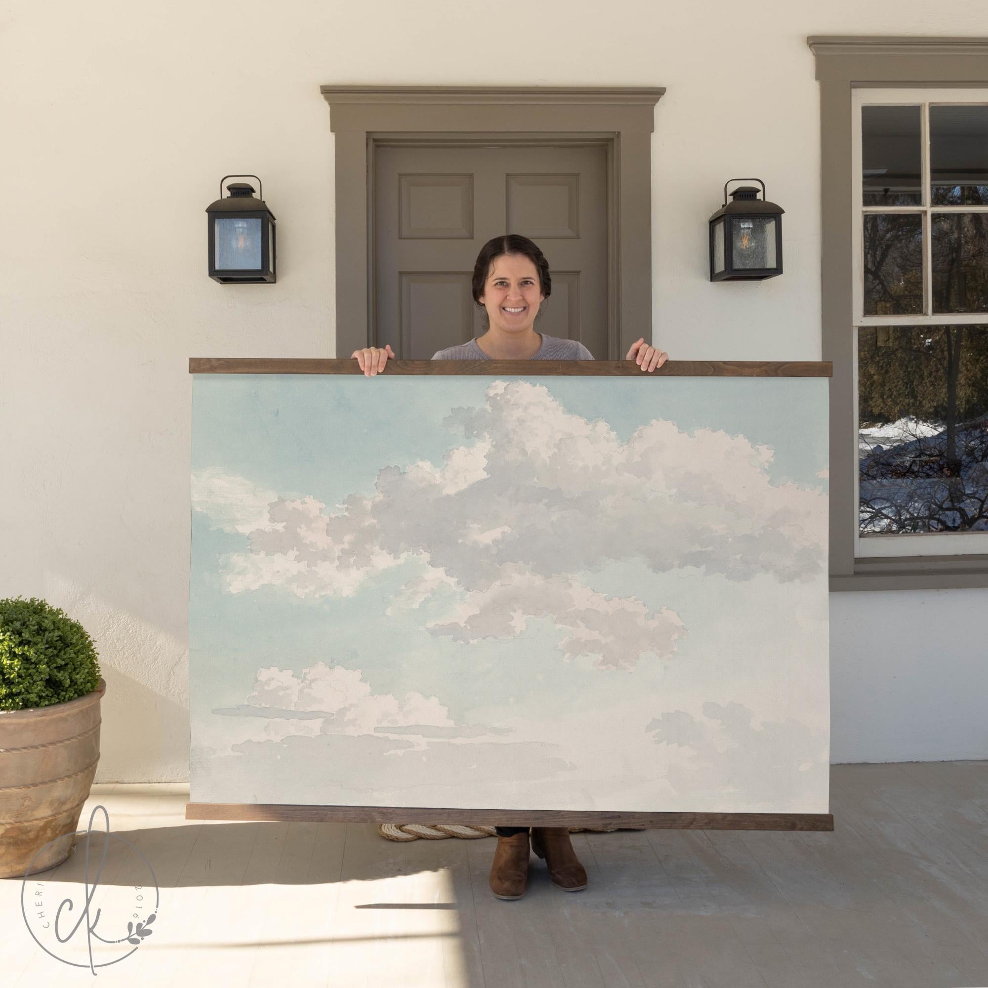 A person holding a large wall art piece featuring a bright blue sky with fluffy white clouds. The background includes a house entrance with a door, wall-mounted lights, and a potted plant.