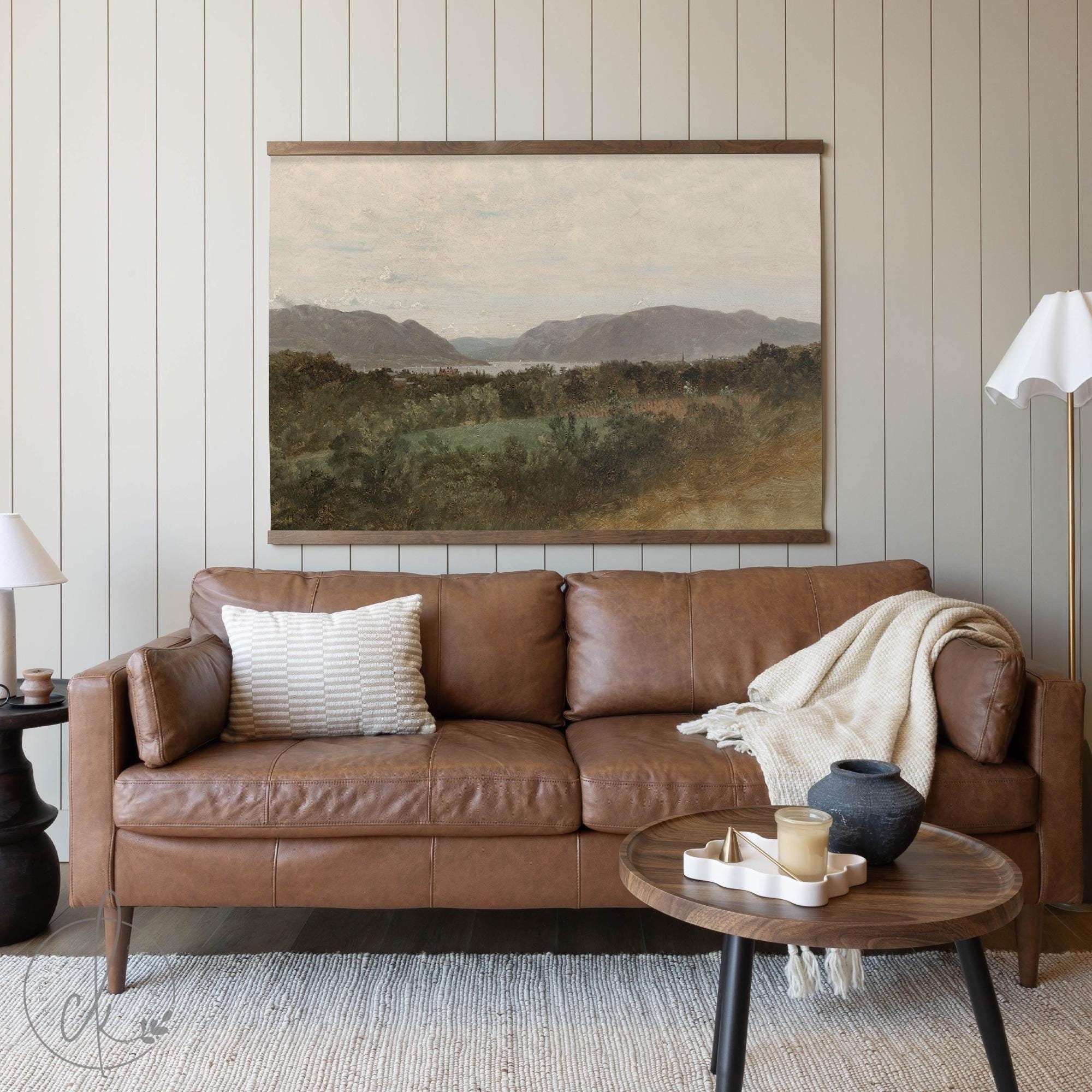 A cozy living room featuring a brown leather sofa with a textured pillow and a draped blanket. A round wooden coffee table with decor sits in front, and a wall-mounted art piece depicting a scenic landscape with hills and greenery hangs above.