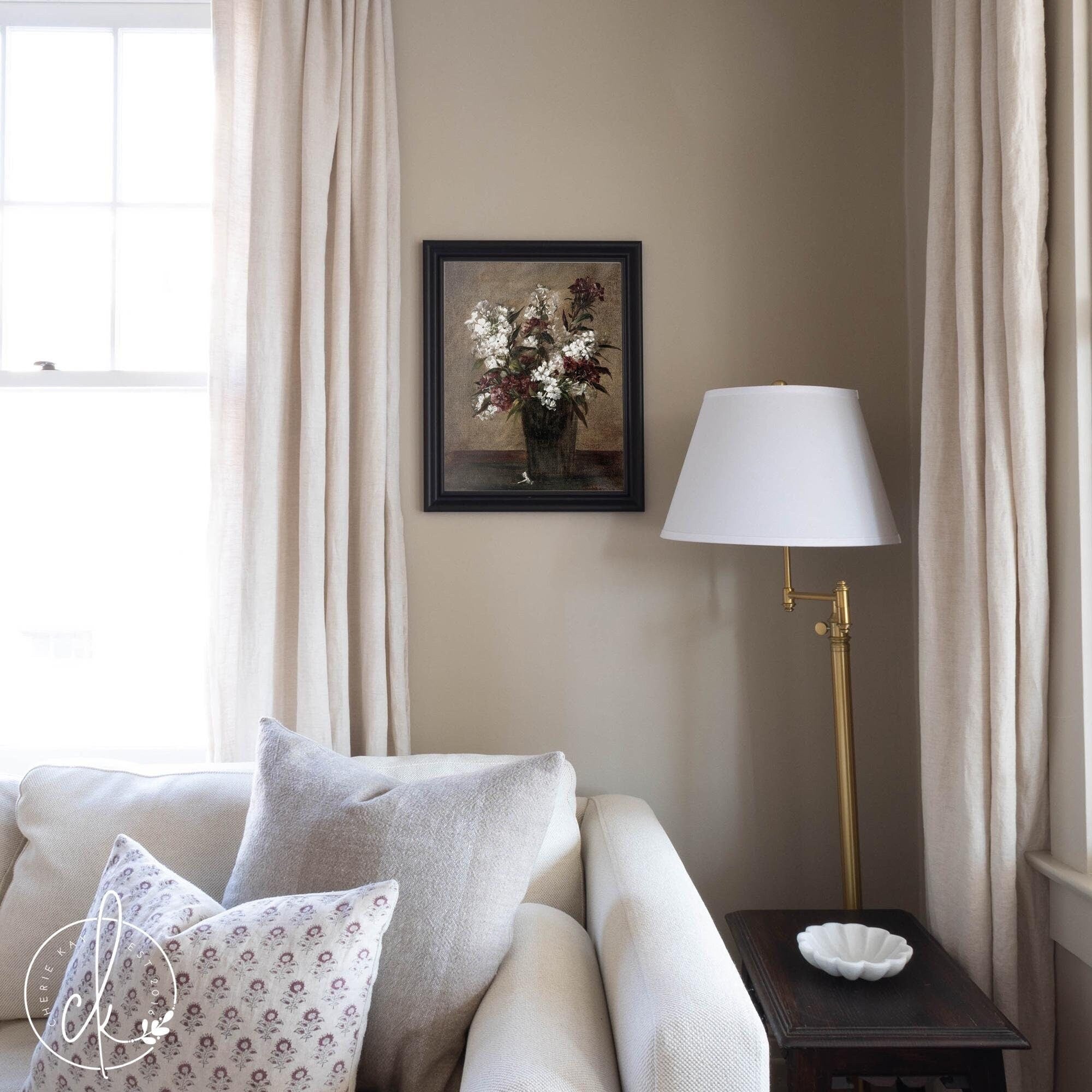 A cozy living room corner with a framed vintage moody floral art print, a brass floor lamp, a wooden side table, and a cream-colored sofa with textured pillows.