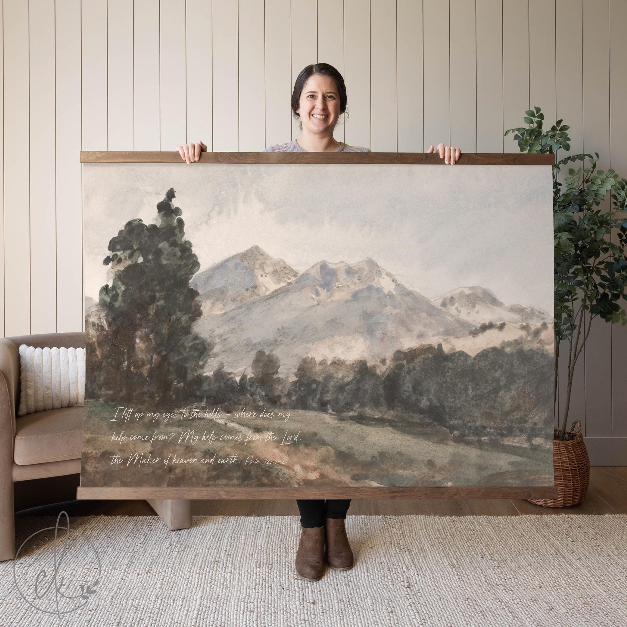 A person holding a wall art piece of a mountain landscape with a verse from Psalm 121:1-2. The background features a neutral interior with a sofa and plant.