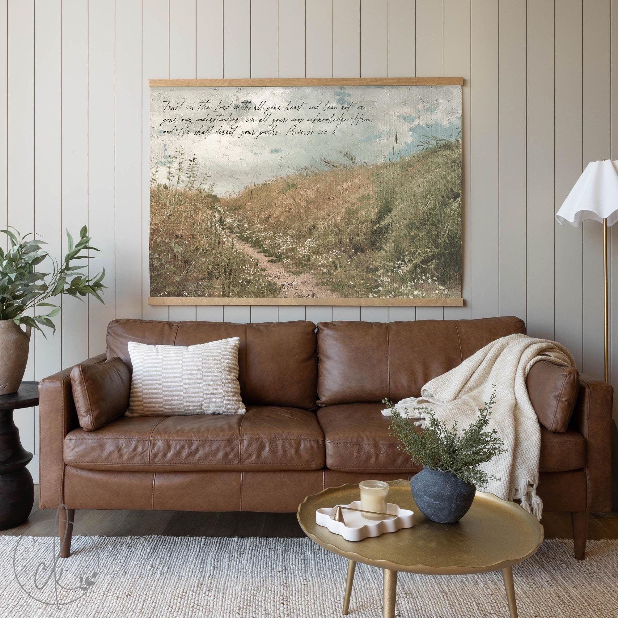 A cozy living room with a brown leather sofa, a textured pillow, and a draped blanket. A gold coffee table with simple decor sits in front. A wall-mounted art piece of a grassy path with a scripture verse from Proverbs 3:5-6 hangs above.