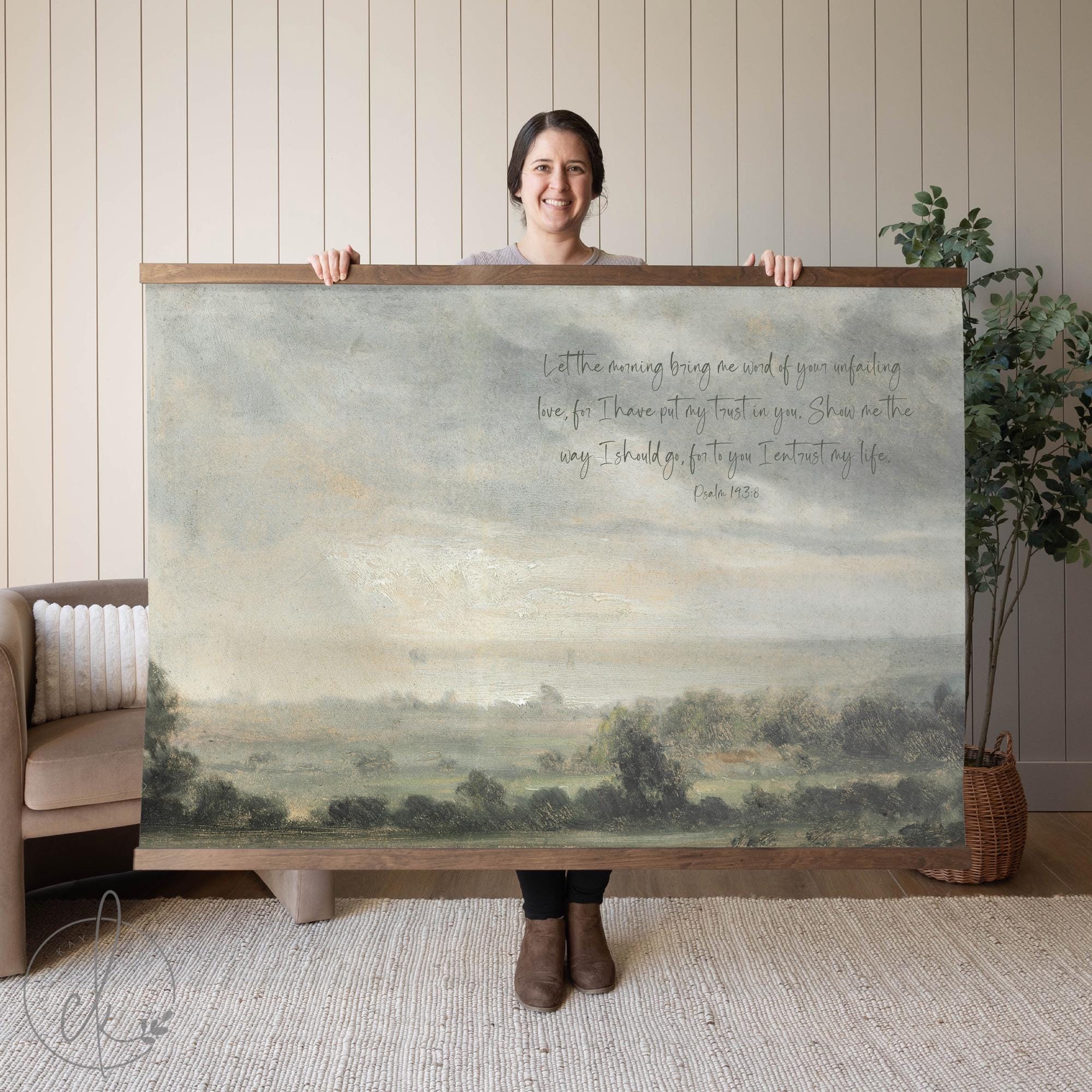 A person holding wall art of a serene landscape with a verse from Psalm 143:8. The background features a sofa and a potted plant.