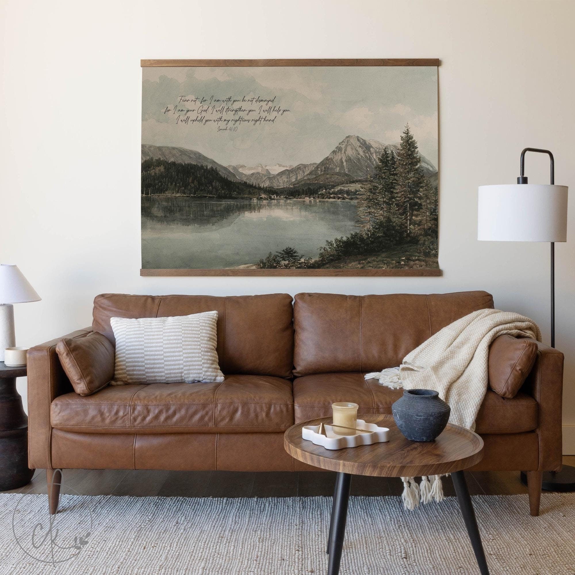 A cozy living room with a brown leather sofa, a textured pillow, and a blanket draped over the arm. A round wooden coffee table with decor sits in front, and a wall-mounted art piece of a serene mountain lake scene with a scripture verse hangs above.