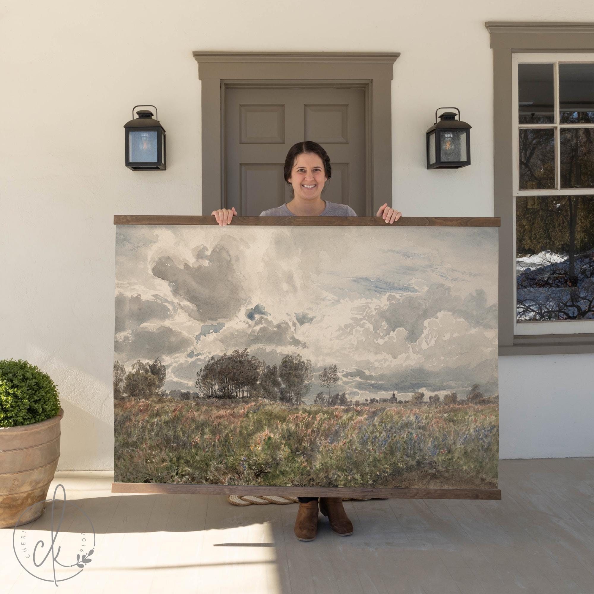 A person standing outside holding a large wall art piece depicting a vibrant field with wildflowers, trees, and a dramatic cloudy sky. The background includes a house entrance with a door, wall-mounted lights, and a potted plant.