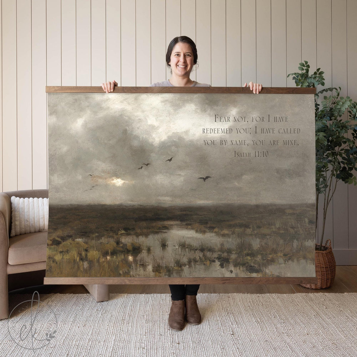 A person holding a large wall art piece featuring a serene marsh landscape with birds and a scripture verse from Isaiah 43:1. The background includes neutral decor, a sofa, and a potted plant.