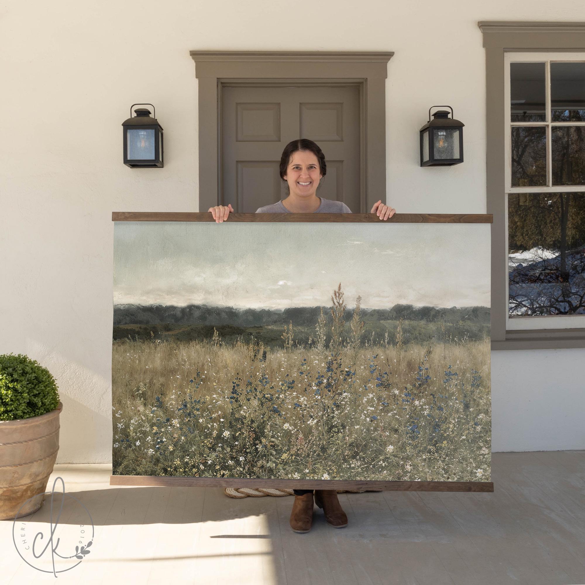 Large framed landscape wall art featuring a serene wildflower meadow with soft neutral tones, held by a woman standing in front of a rustic home entrance.