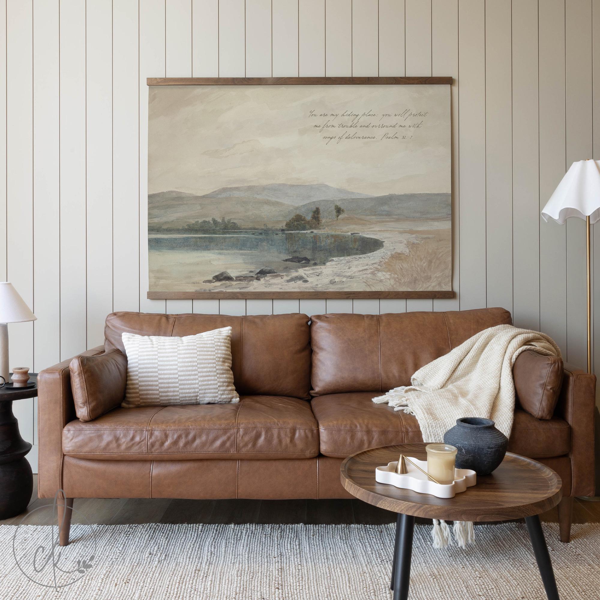 A cozy living room with a brown leather sofa, a wooden coffee table, and a large framed landscape artwork with a Bible verse on the wall.