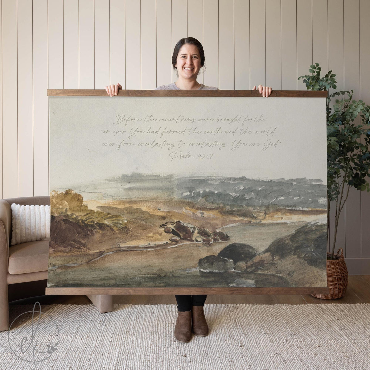 A person holding a large canvas tapestry wall art piece of a serene landscape with a scripture verse from Psalm 90:2. The background features a neutral interior with a sofa and a potted plant.