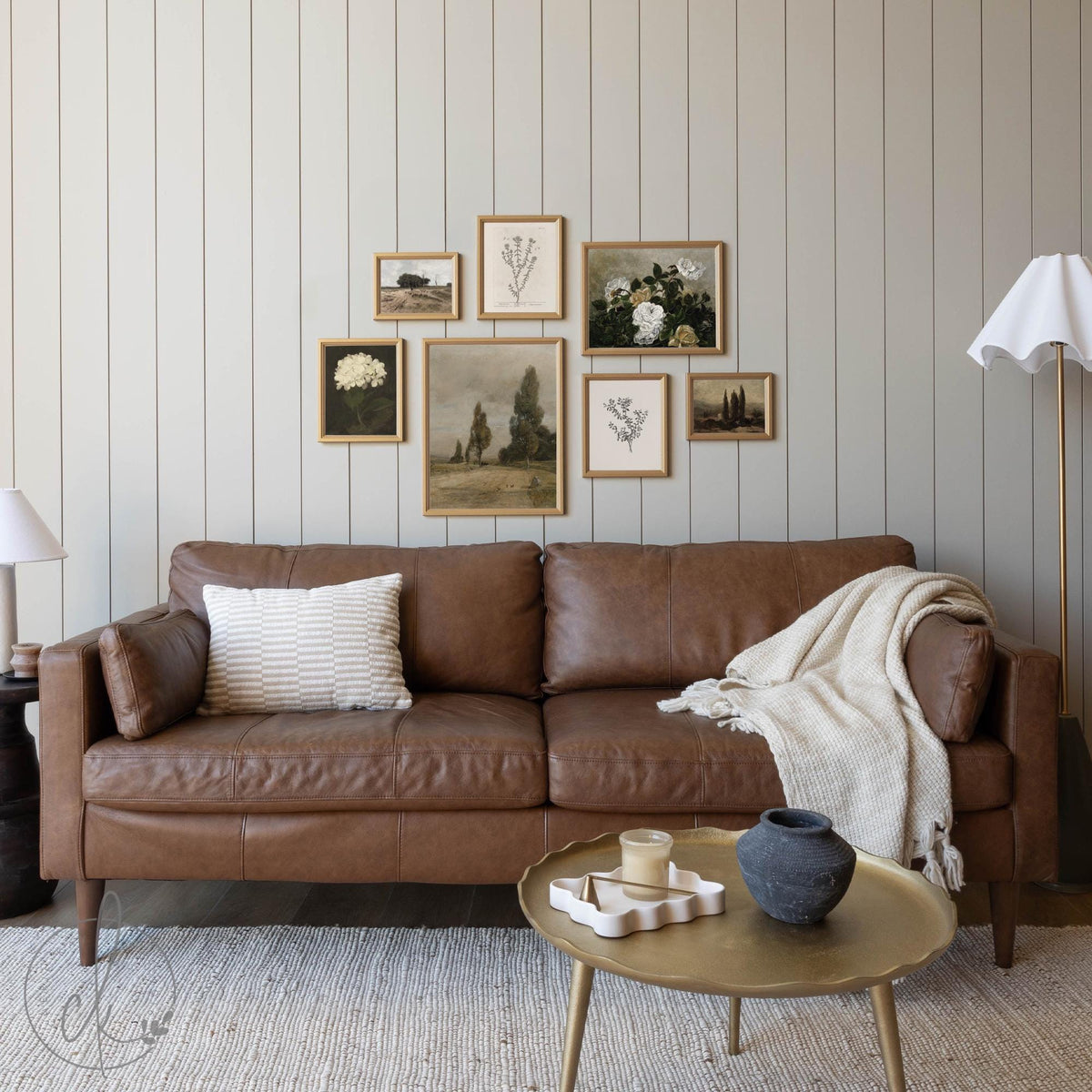 Framed vintage gallery wall art set featuring a mix of botanical, floral, and landscape prints in warm wood frames, arranged above a brown leather sofa in a neutral-toned living room.