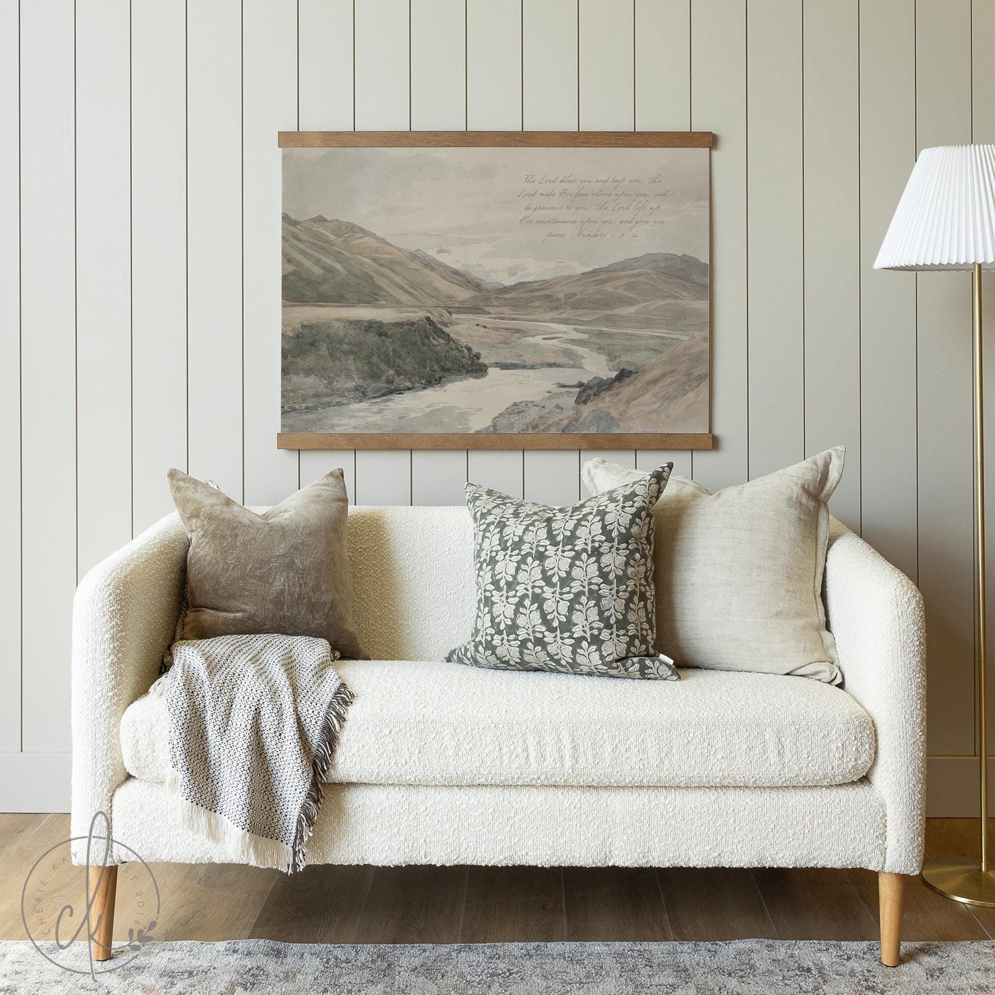 A cozy living room with a white boucle sofa, neutral throw pillows, a textured blanket, and a vintage landscape wall tapestry of mountains and a river.