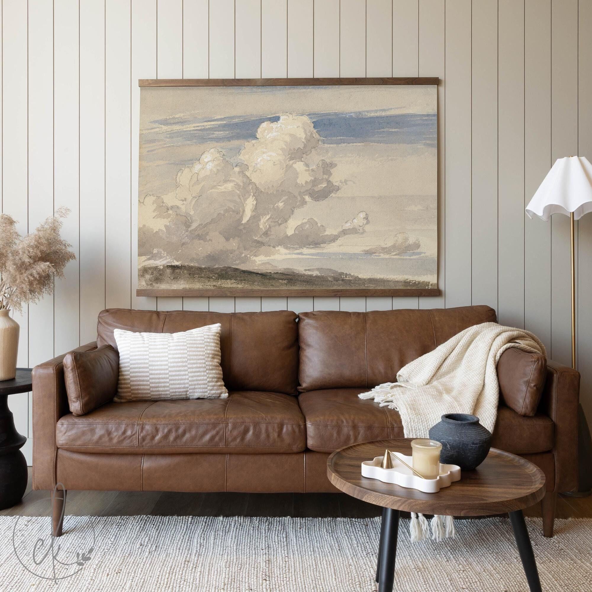 Framed vintage landscape tapestry featuring a cloud-filled sky, hanging above a brown leather couch in a cozy, neutral-toned living room.