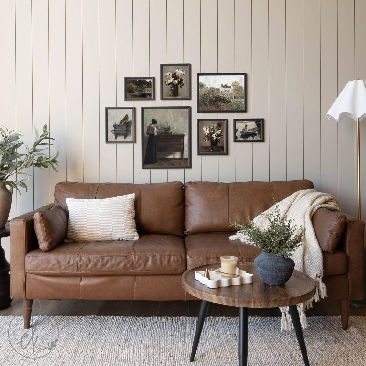 Framed vintage gallery wall art set displayed above a brown leather sofa in a cozy living room. The arrangement features antique-inspired prints with floral, landscape, and portrait themes, creating a timeless and elegant aesthetic. Neutral tones complement the rustic and classic decor.