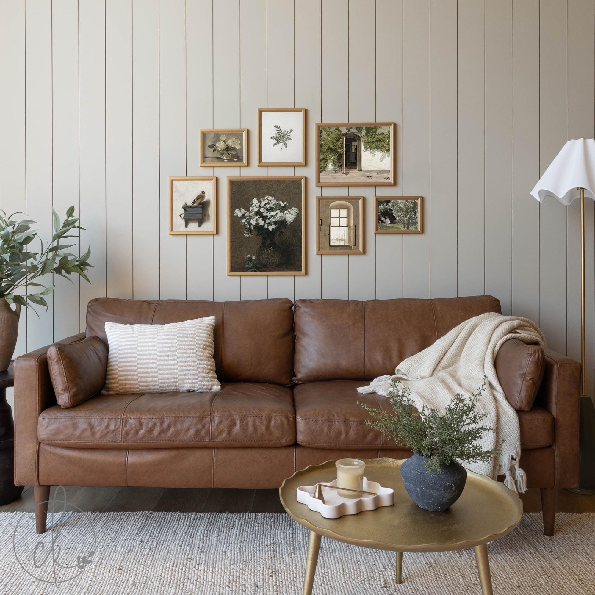 Framed vintage gallery wall art set displayed above a brown leather sofa, featuring a curated mix of botanical prints, European-inspired architecture, and still-life paintings in warm, neutral tones, creating a cozy and rustic living room aesthetic.