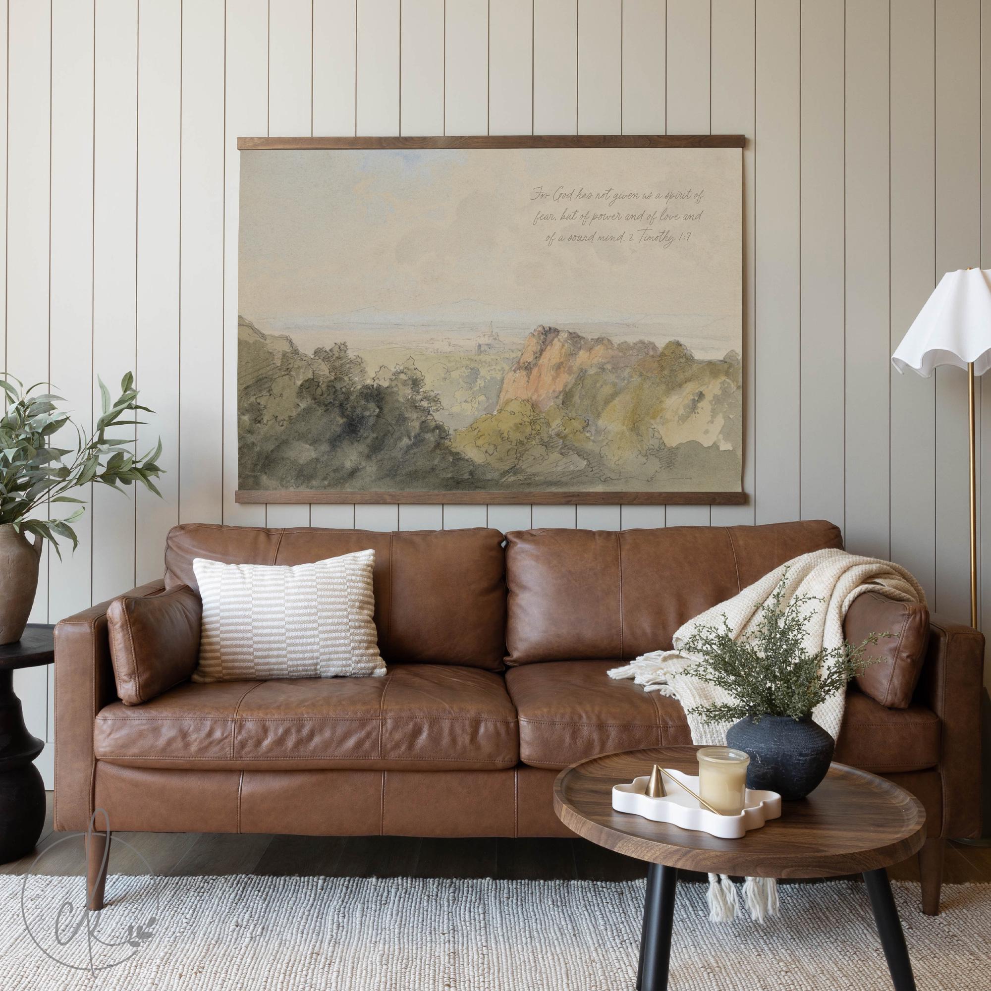 Cozy living room with a brown leather sofa, neutral decor, and a large vintage landscape tapestry hanging above the couch. The tapestry features a serene countryside scene with soft muted tones and an elegant wooden hanger, complementing the rustic and farmhouse aesthetic.