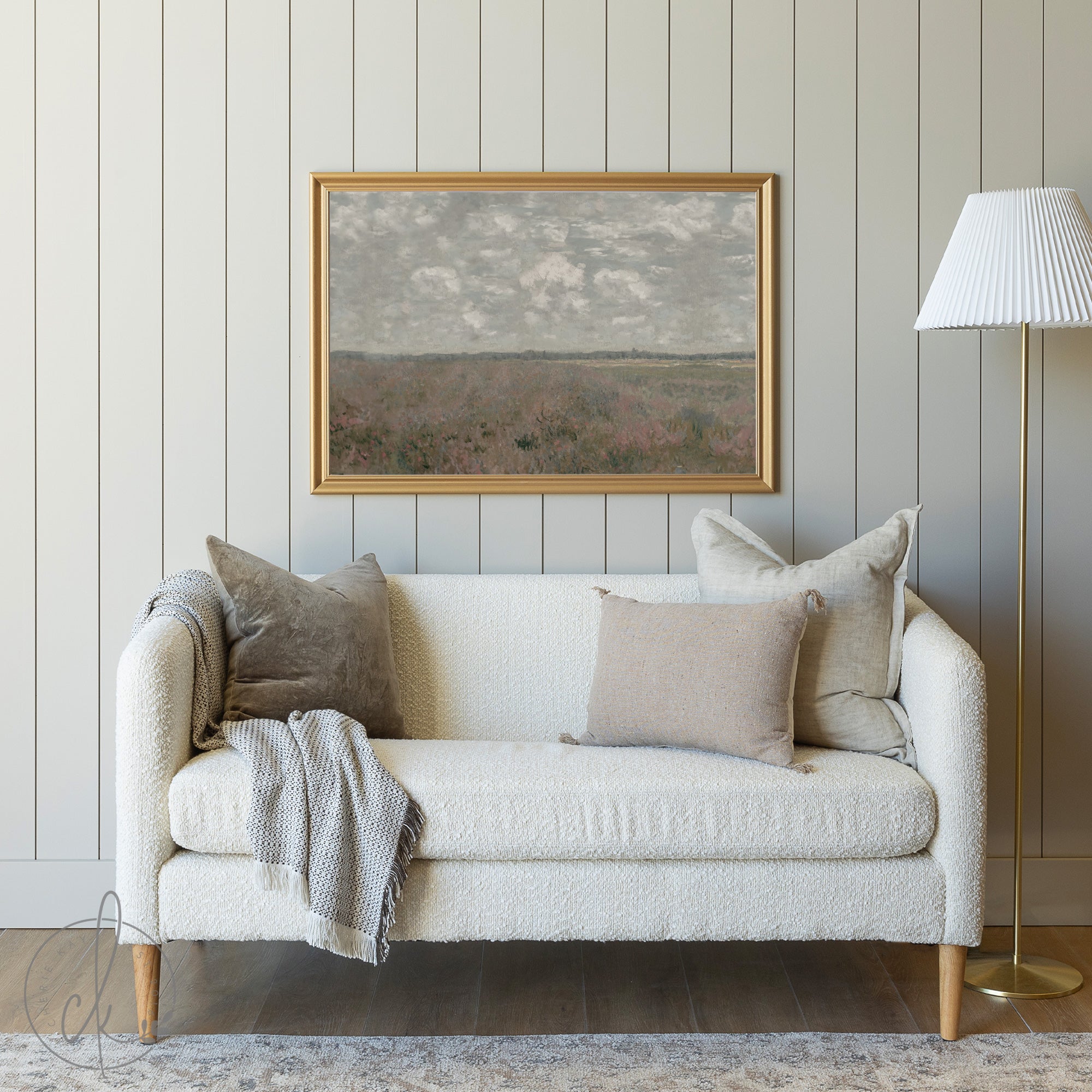 A cozy living room with a cream sofa, neutral pillows, a gray blanket, and a framed painting of a cloudy sky over a serene landscape on a muted paneled wall. A gold floor lamp with a white shade stands nearby.