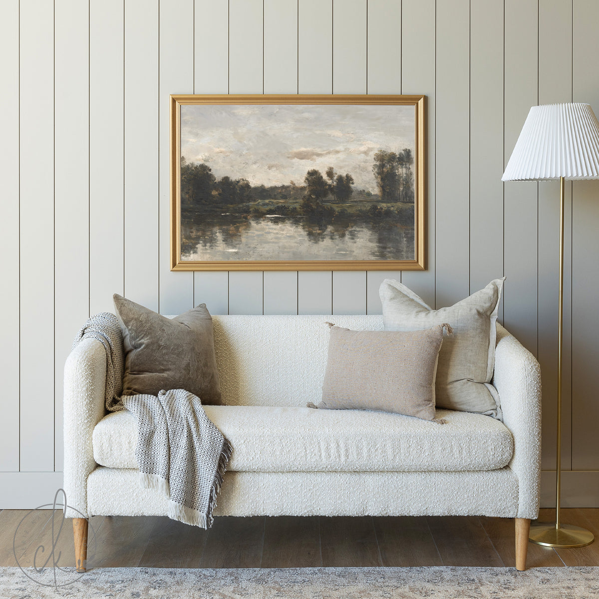 
A cozy living room featuring a cream sofa with neutral throw pillows and a gray blanket draped over one arm. Above the sofa, a framed painting of a tranquil riverscape with trees and reflections hangs on a muted paneled wall. A gold floor lamp with a white pleated shade stands nearby.