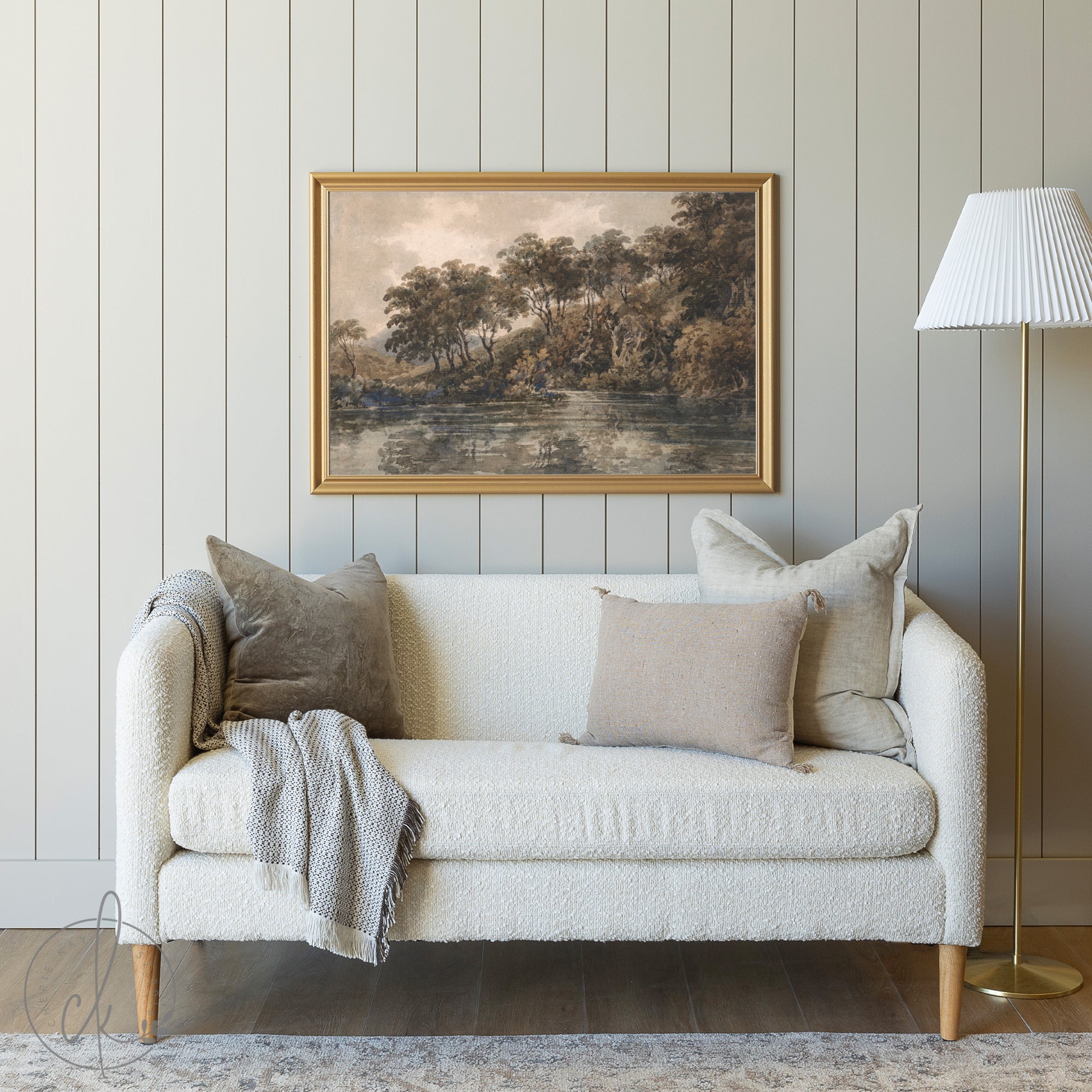 A cozy living room with a cream sofa, neutral throw pillows, and a gray blanket, set against a paneled wall. Above the sofa, a framed painting of a serene riverside landscape with trees and reflections hangs. A gold floor lamp with a white pleated shade stands to the side.