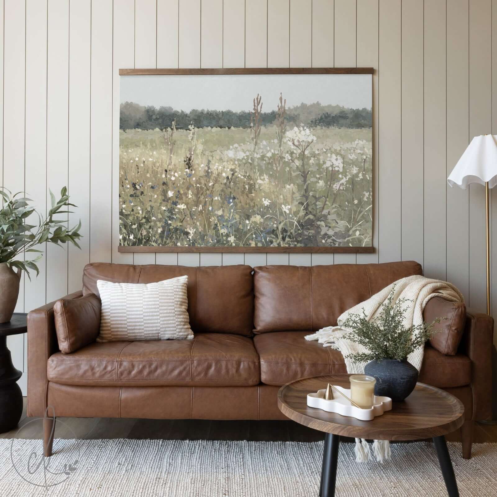 A cozy living room with a brown leather sofa, a wooden coffee table, and a potted plant, featuring a large painting of a floral field on the wall.