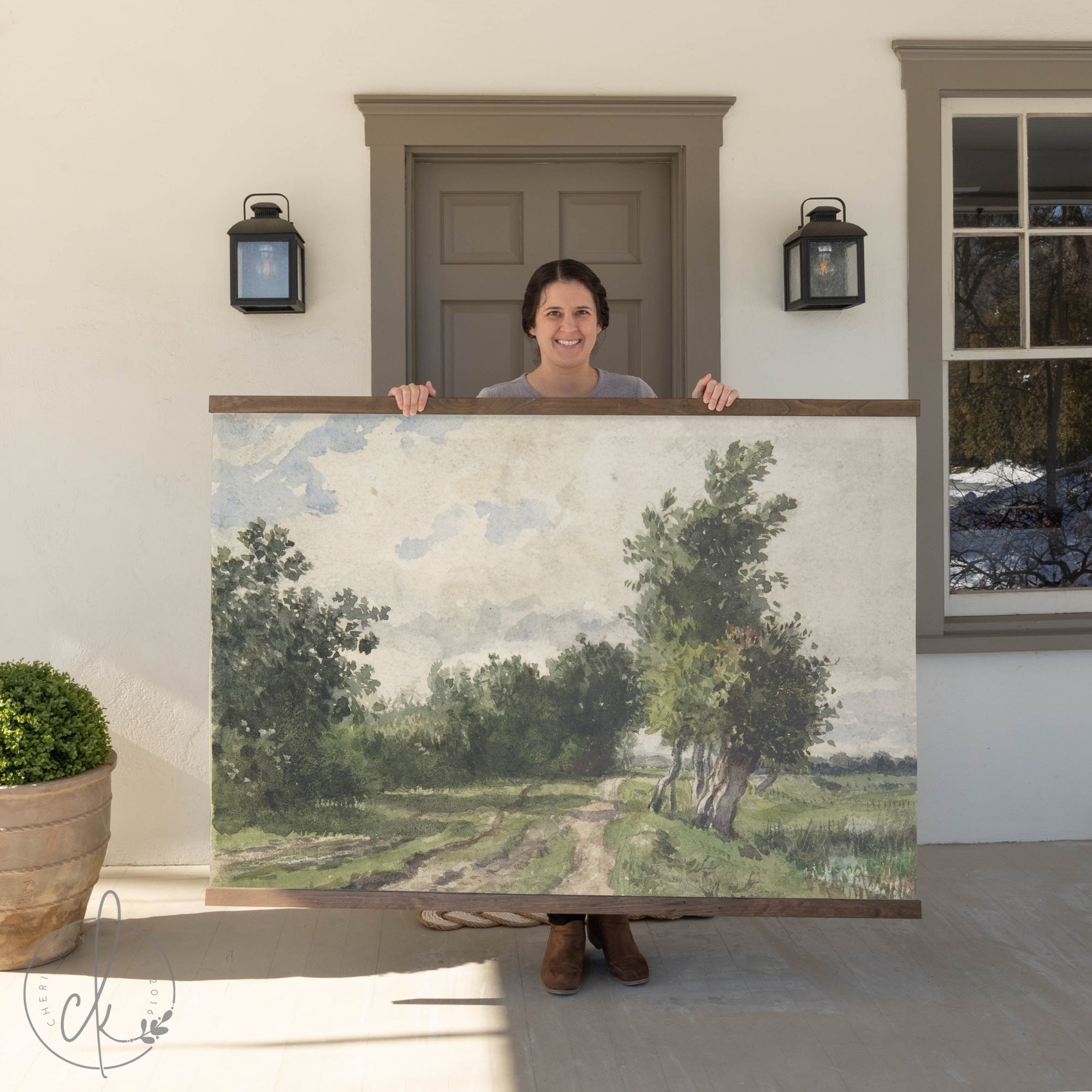 A woman stands on a porch holding a large framed landscape painting featuring a rural path lined with trees under a partly cloudy sky.