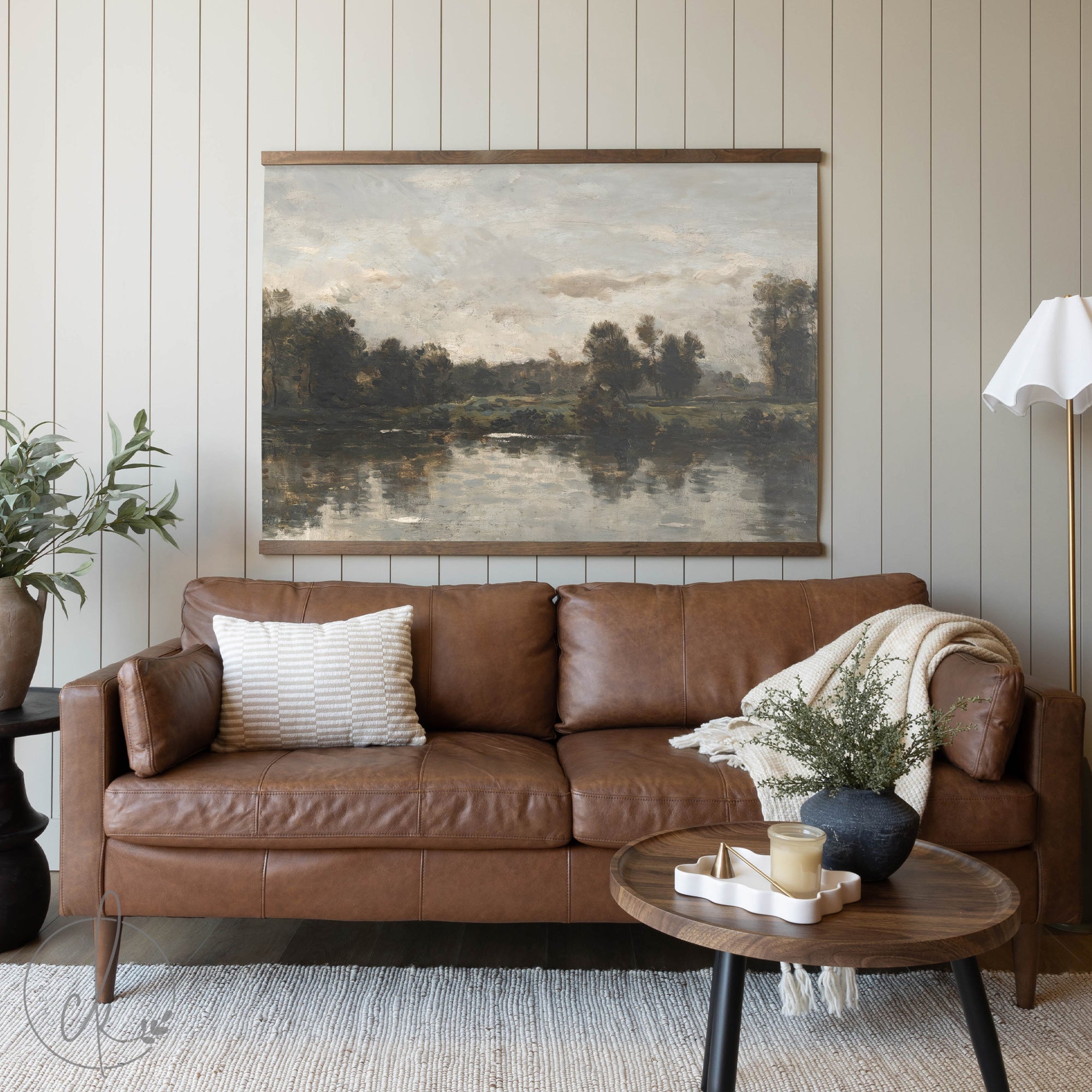 A cozy living room with a brown leather sofa, a wooden coffee table, and a potted plant, featuring a large painting of a serene river landscape on the wall.