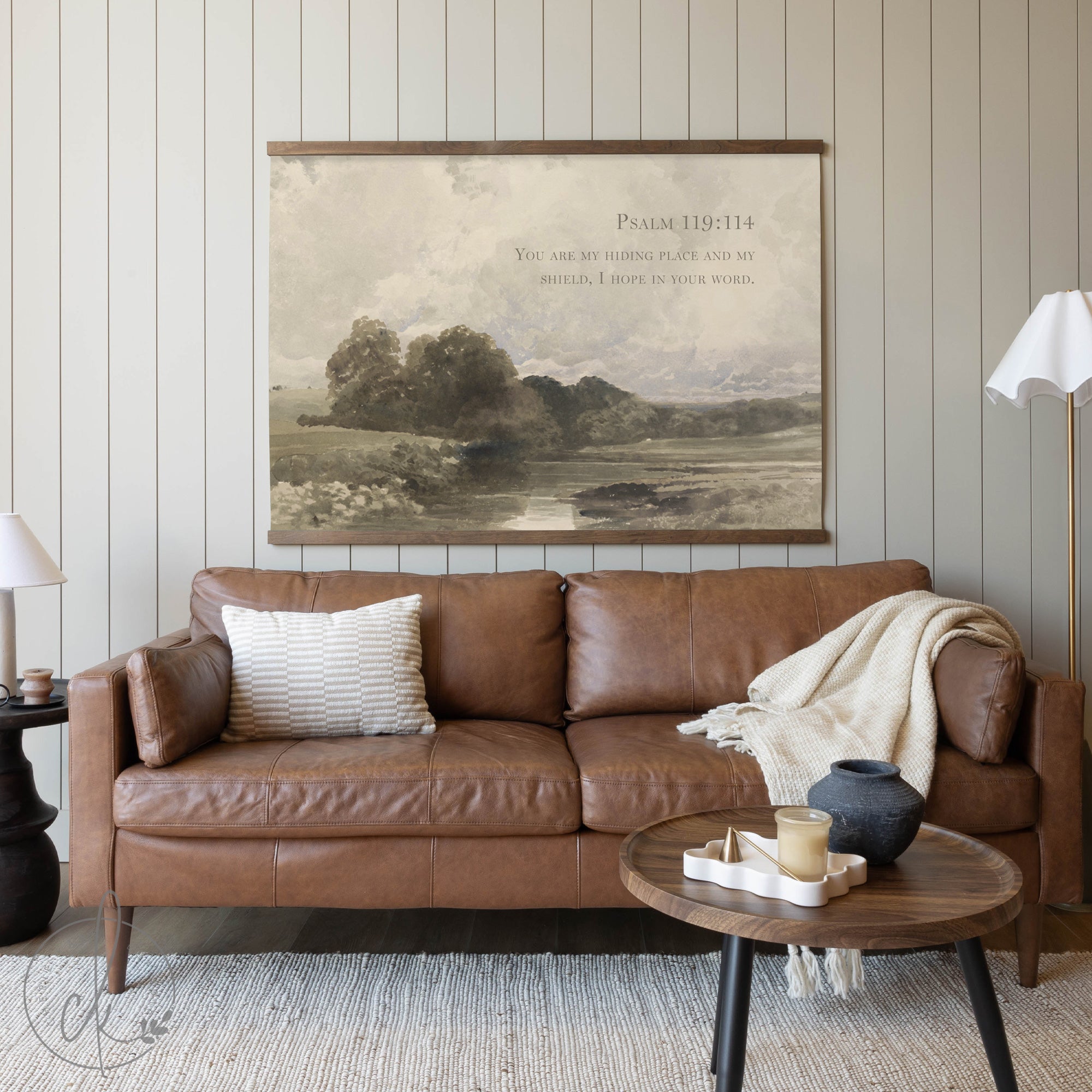 A cozy living room with a brown leather sofa, a wooden coffee table, and a potted plant, featuring a large painting with the Psalm 119:114 verse and a landscape on the wall.