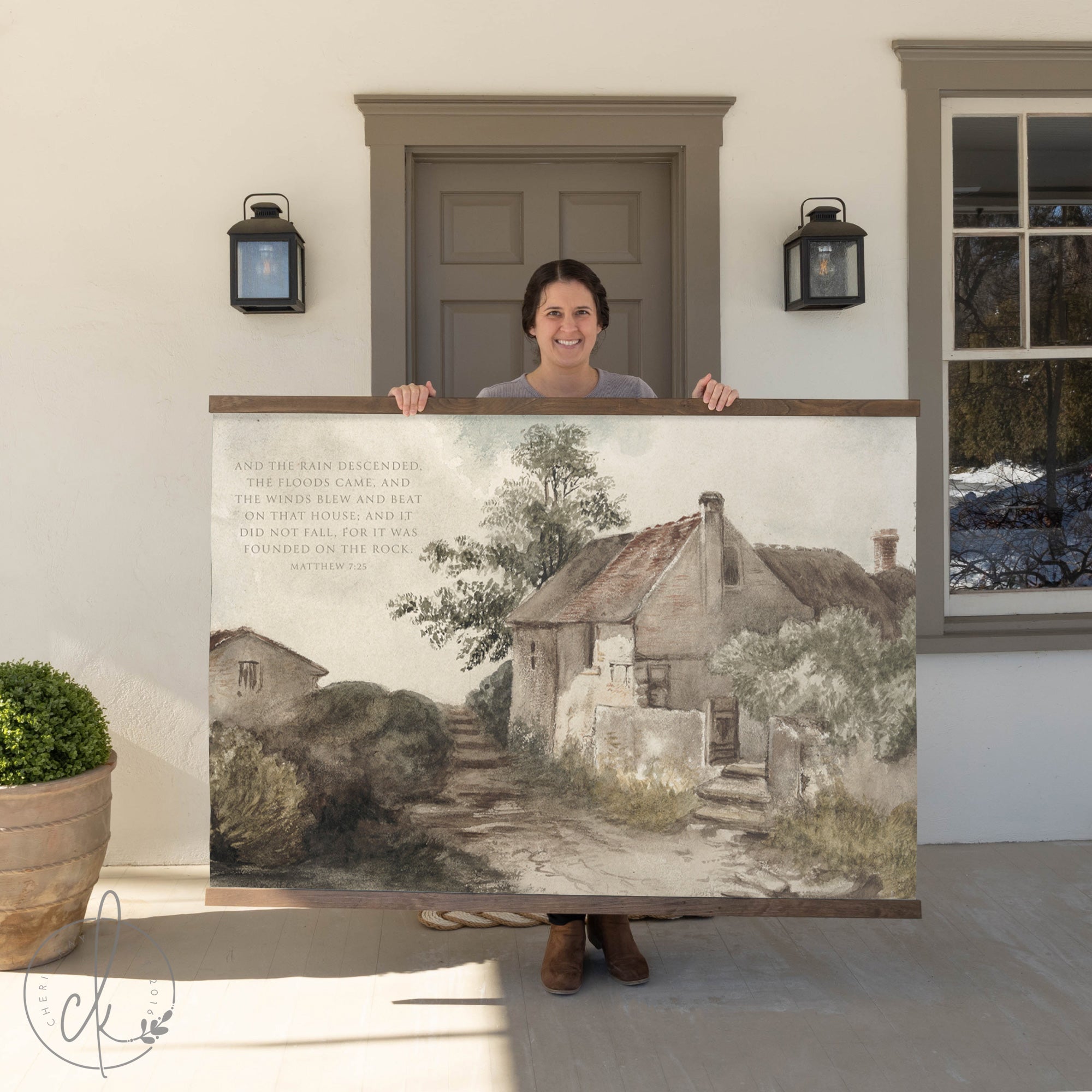 A woman holding a large painting of a house and pathway, with a quote from Matthew 7:25 written on it, standing in front of a doorway.