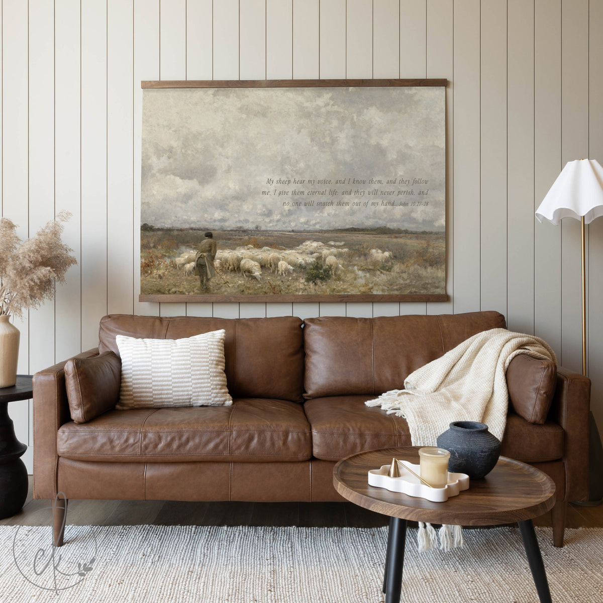 A cozy living room featuring a brown leather sofa, a wooden coffee table, and a pastoral painting on the wall depicting a shepherd with a flock of sheep, accompanied by a Bible verse from John 10:27-28.