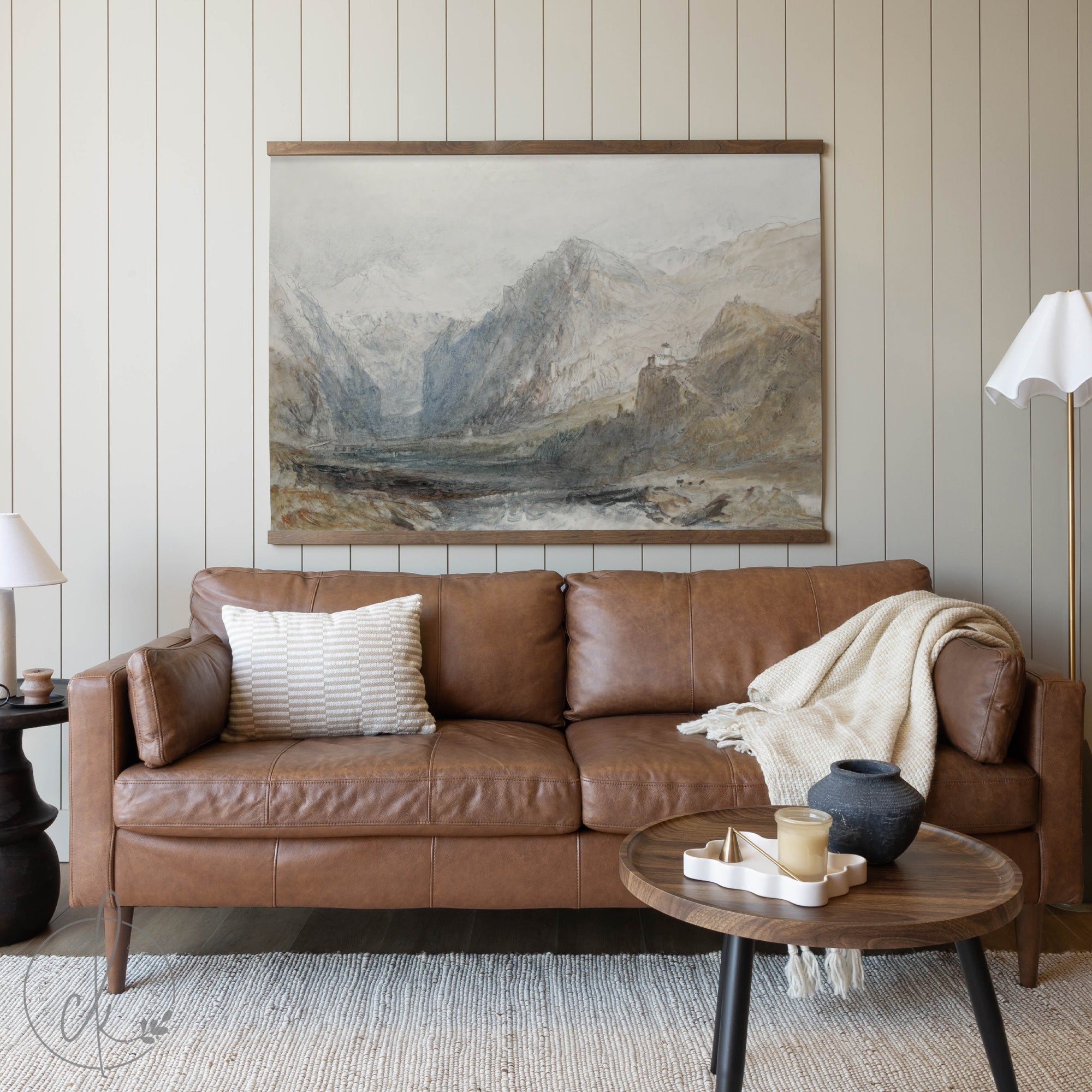 A cozy living room with a brown leather sofa, a wooden coffee table, and a potted plant, featuring a large painting of a mountain landscape on the wall.