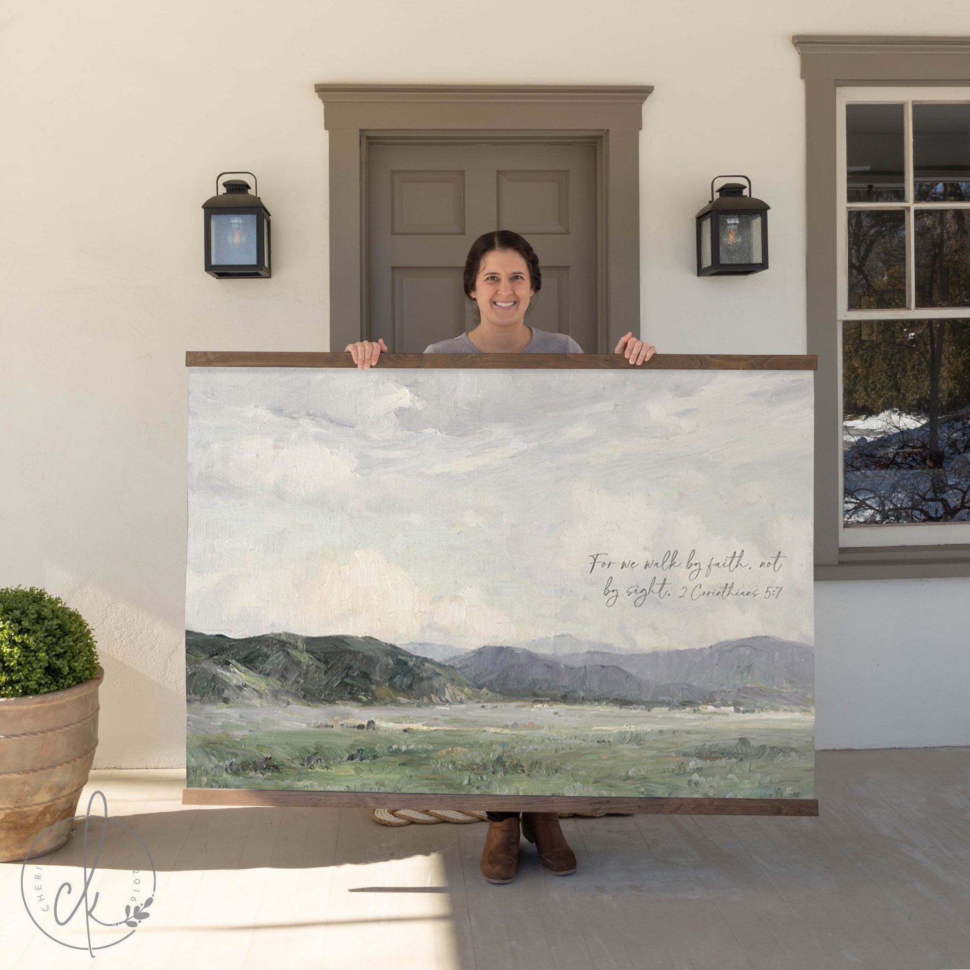 A woman stands on a porch holding a large framed landscape painting featuring rolling green hills and a cloudy sky, with the Bible verse "For we walk by faith, not by sight. 2 Corinthians 5:7" written on it.