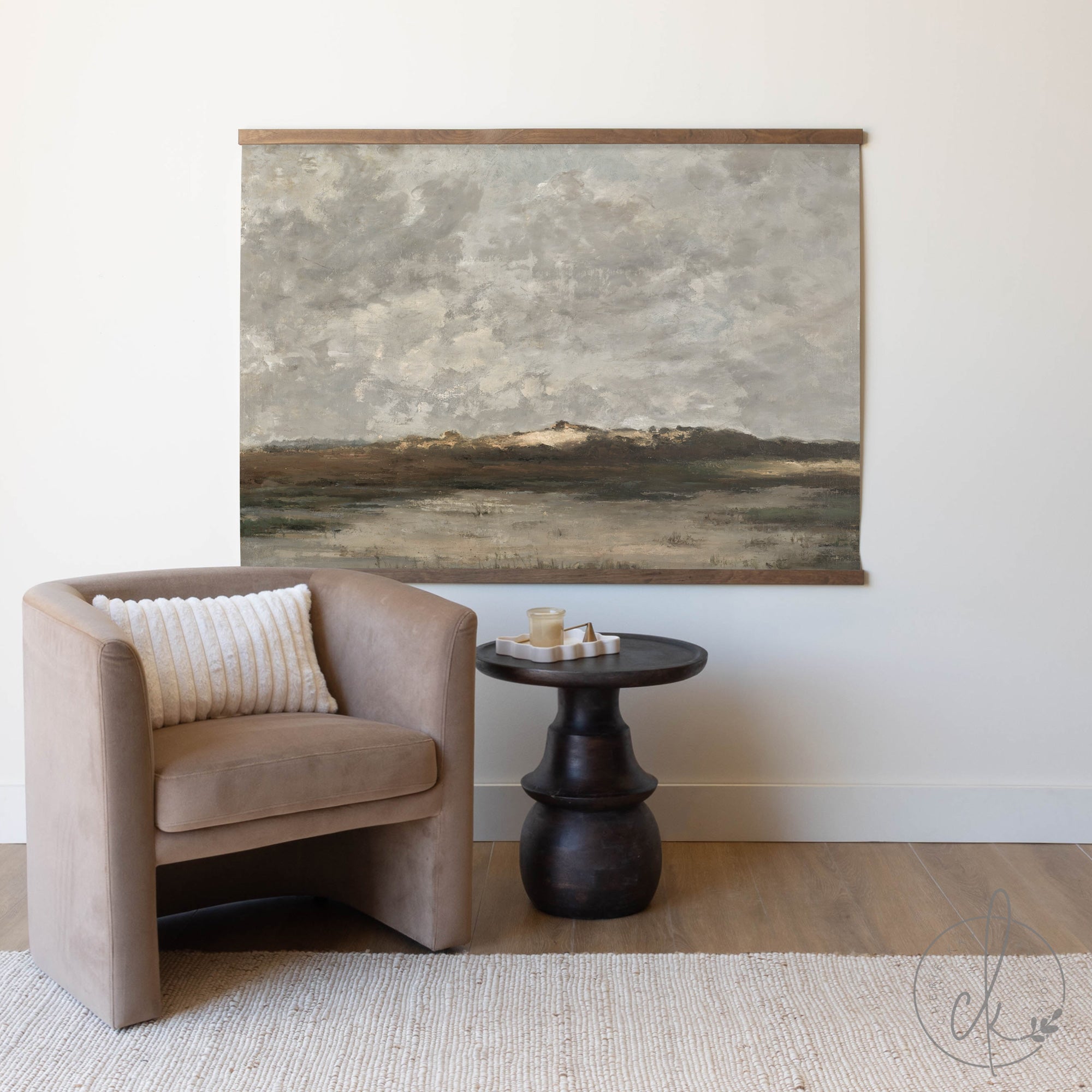 A minimalist and cozy corner featuring a beige accent chair with a textured white pillow, a dark wooden side table with a candle tray, and a large framed abstract landscape painting on the wall. The neutral color palette and soft textures create a calming atmosphere.